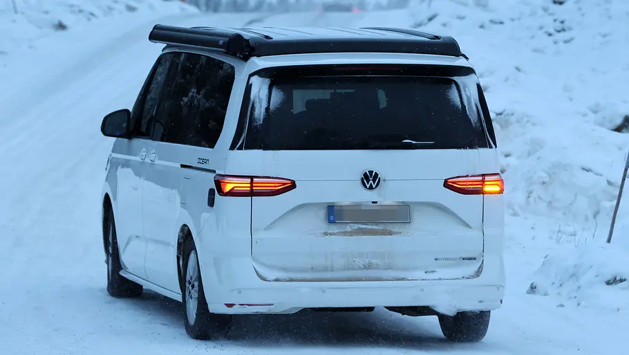 Ein Erlkönig des VW California Facelift auf einer Erprobungsfahrt auf verschneiter Straße in Nordschweden von schräg hinten links.