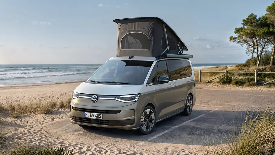 Das VW California Facelift statisch von schräg vorne links mit geöffnetem Aufstelldach vor einem Strand.