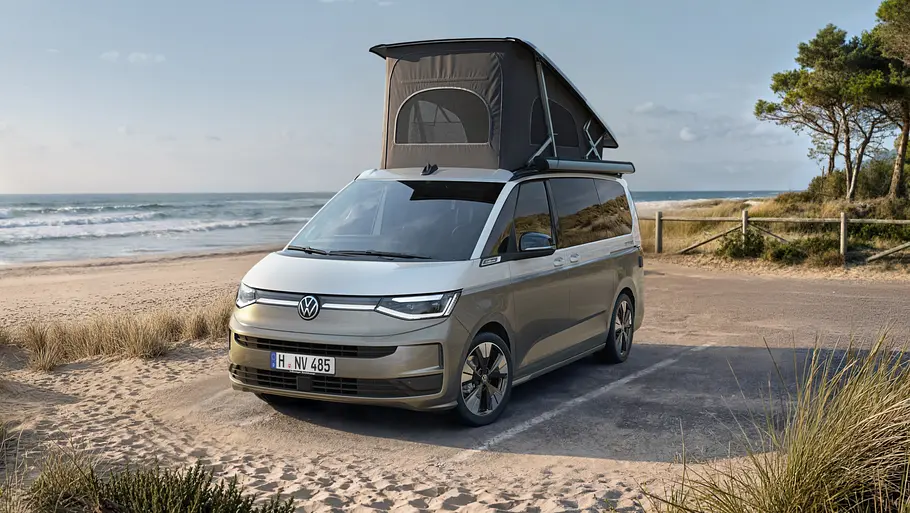 VW California Facelift Der VW California statisch von schräg vorne links mit geöffnetem Aufstelldach neben einem Strand.