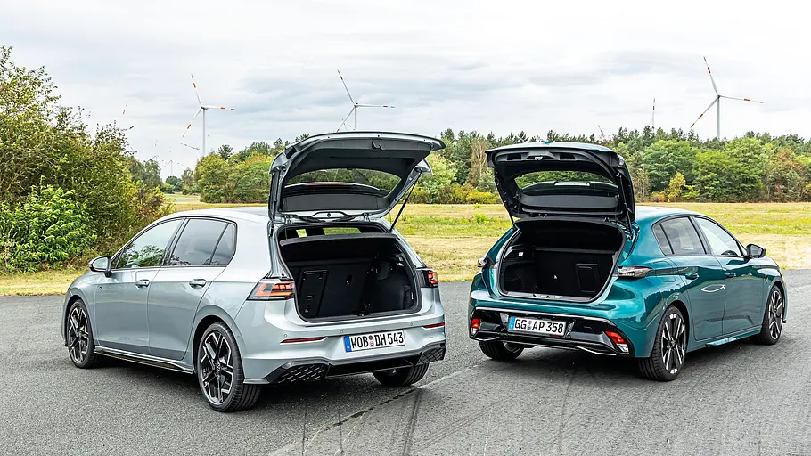 Ein silberner VW Golf und ein türkisfarbener Peugeot 308 stehen mit offenen Heckklappen nebeneinander. Dahinter sind Bäume und Windräder zu sehen.