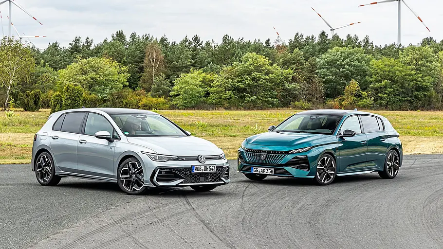 Ein silberner VW Golf und ein türkisfarbener Peugeot 308 stehen auf Asphalt. Dahinter sind Wald und Windräder unter bewölktem Himmel.