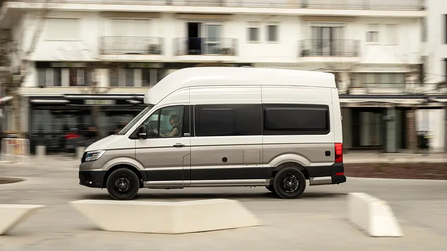 VW Grand California Facelift Fahrbericht Das VW Grand California Facelift fahrend im Profil von links vor einer Innenstadt-Szenerie.