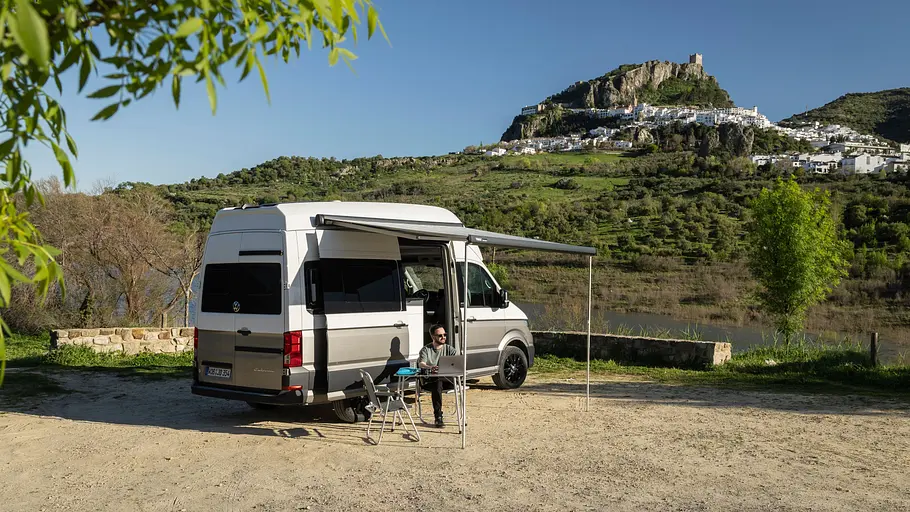 VW Grand California Facelift Fahrbericht Das VW Grand California Facelift statisch von schräg hinten rechts mit ausgefahrener Markise auf einem Schotterparkplatz. Unter der Markise sitzt eine Person am Falttisch auf einem Campingstuhl, im Hintergrund ist die Stadt Zahara de la Sierra zu sehen.