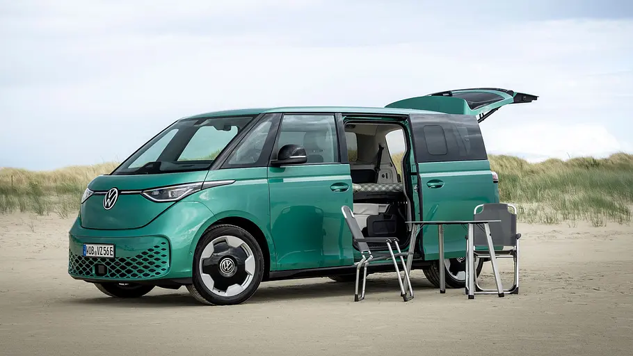 VW ID. Buzz mit Gute-Nacht-Paket Der VW ID. Buzz mit Gute-Nacht-Paket statisch von schräg vorne links am Strand. Schiebetüren und Heckklappe sind geöffnet, vor dem Fahrzeug steht ein Campingstuhl- und -tisch-Set.