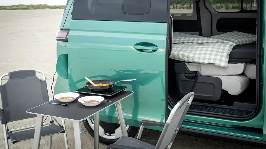 VW ID. Buzz mit Gute-Nacht-Paket Campingmöbel an der geöffneten rechten Schiebetüre des VW ID. Buzz mit Gute-Nacht-Paket. Durch die Schiebetüre ist das Bett zu sehen.