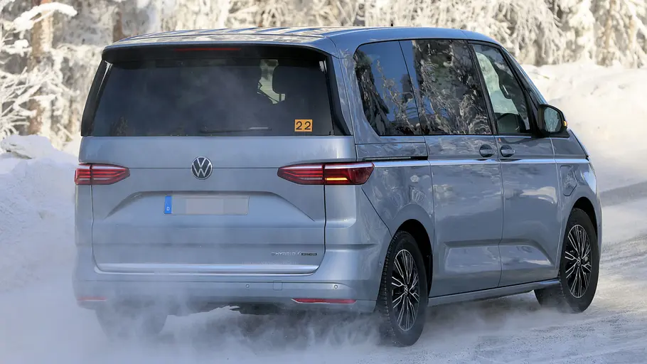 Das VW Multivan Facelift (2026) fährt auf einer verschneiten Straße, von hinten rechts fotografiert.