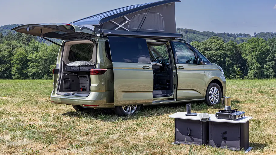 VW-Neuheiten auf dem Caravan Salon 2026 Der VW California Beach statisch von schräg hinten rechts mit geöffneter Heckklappe, rechter Schiebetüre und Aufstelldach auf einer Wiese. Vor dem Fahrzeug steht die Heckküche von Stuff Bubble.