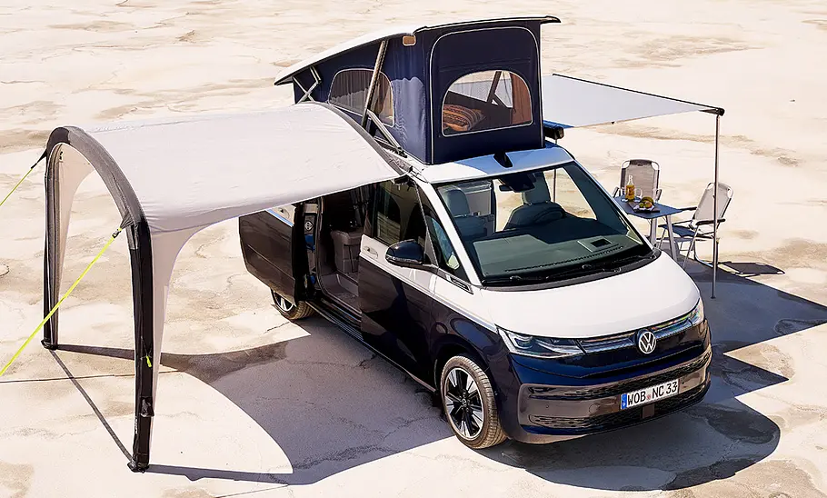 VW California (2024) Das Bild zeigt den VW California (2024) mit einem Vorzelt an der rechten und einer Markise an der linken Fahrzeugseite. Unter der Markise stehen Campingstühle. Das Aufstelldach ist geöffnet.