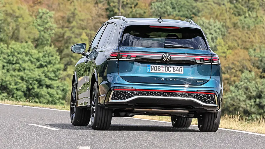 Toyota RAV4 und VW Tiguan im Vergleich Ein blauer VW Tiguan ist schräg von hinten auf einer asphaltierten Straße zu sehen. Im Hintergrund stehen dichte Bäume.