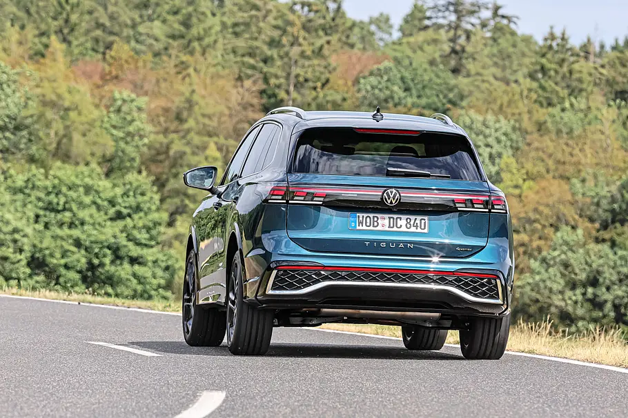 Vergleichstest VW Tiguan gegen Toyota RAV4 Ein blauer VW Tiguan ist schräg von hinten auf einer asphaltierten Straße zu sehen. Im Hintergrund stehen dichte Bäume.