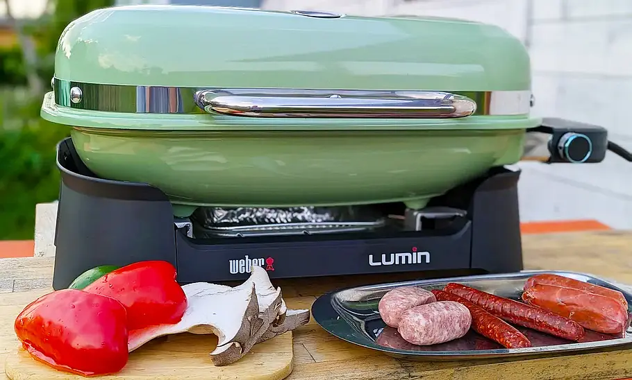 Weber Elektro-Tischgrill Lumin Der Weber Elektro-Tischgrill Lumin von vorne fotografiert. Auf dem Tisch ist zudem ein Tablett mit verschiedenen rohen Wurstsorten und ein Brett mit Paprika und Pilzen abgestellt.