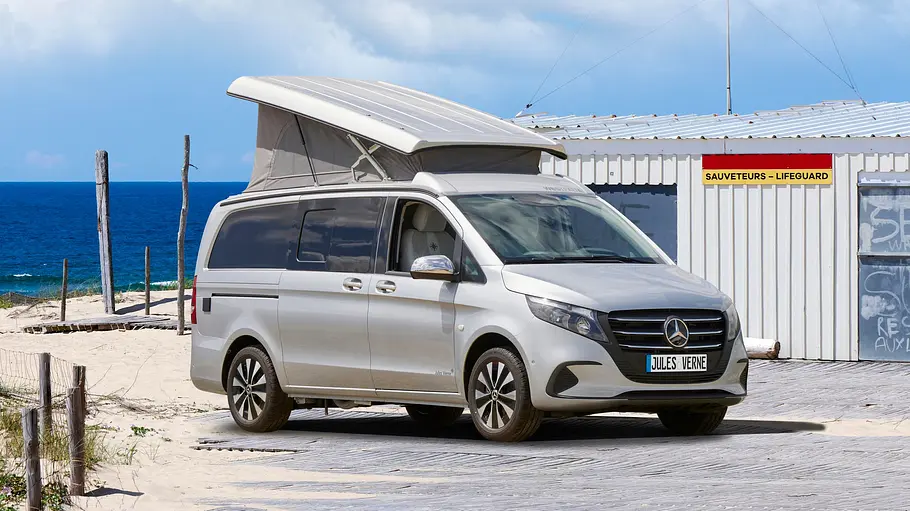 Westfalia Jules Verne Die Außenaufnahme zeigt den Westfalia Jules Verne mit aufgestelltem Aufstelldach von schräg seitlich vor einer Strandkulisse.