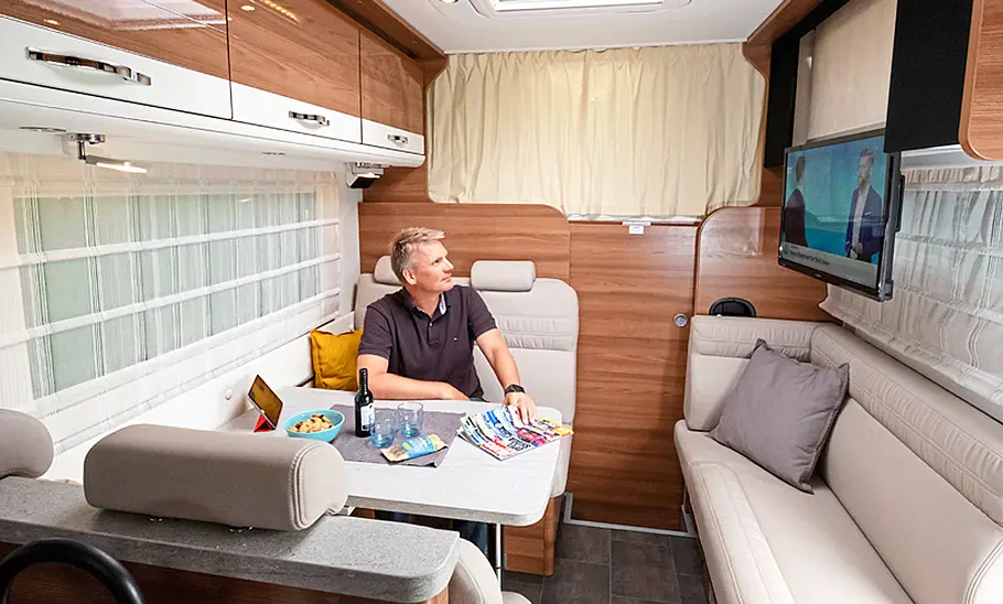 Das Bild zeigt die Sitzgruppe des Dethleffs Globetrotter XXL A mit einer Personen auf einem der Sitze. Die Tür zum Cockpit ist geschlossen, der Fernseher über dem Einzelsofa auf der rechten Fahrzeugseite ist heruntergefahren.
