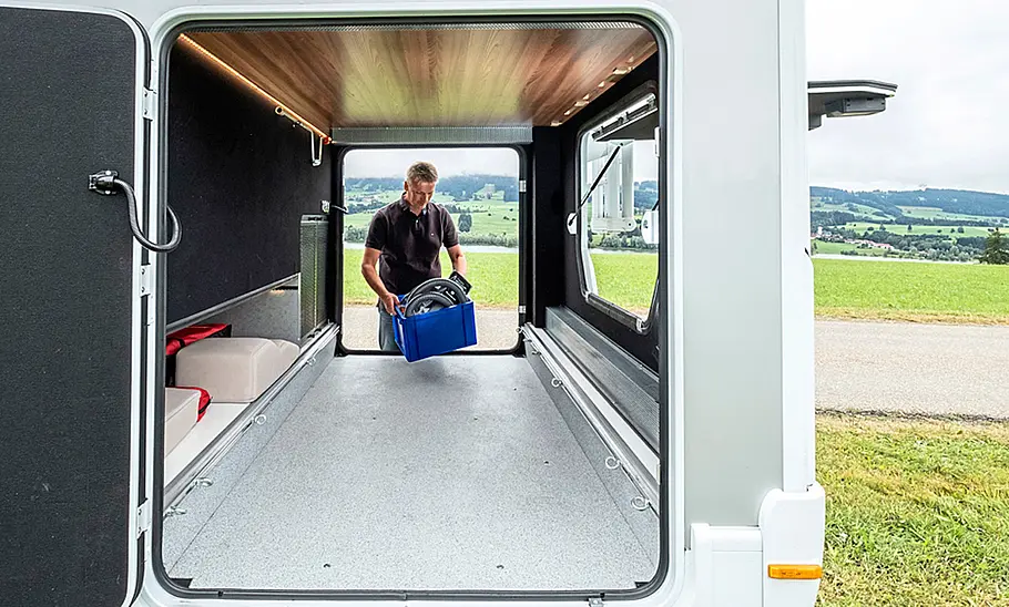 Das Bild zeigt die Heckgarage des Dethleffs Globetrotter XXL A mit geöffneten Klappen zu drei Seiten und einer Person, die Gepäck einlädt.